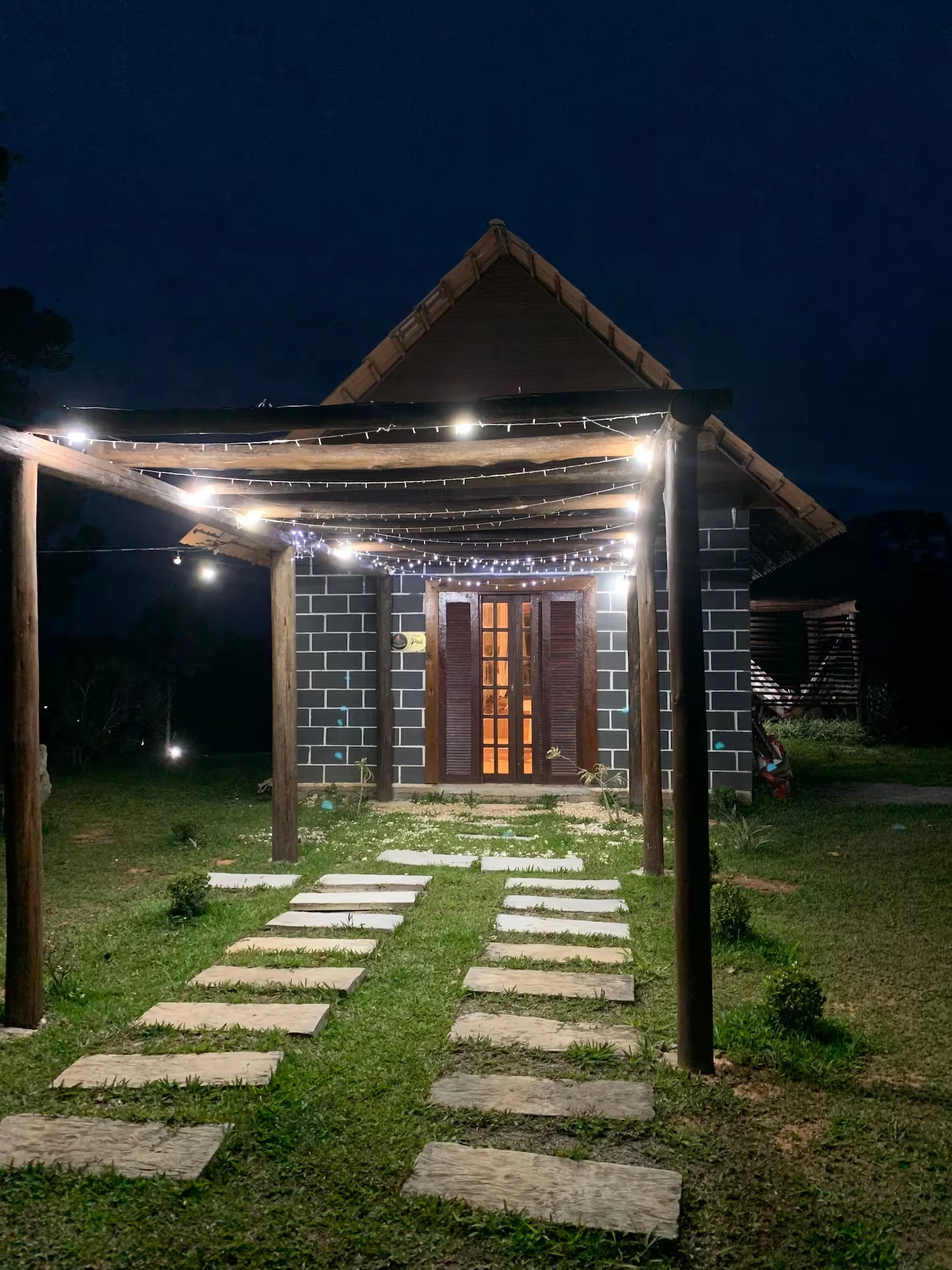 Chalé iluminado à noite, com pergolado de madeira, luzinhas e caminho de pedras até a porta