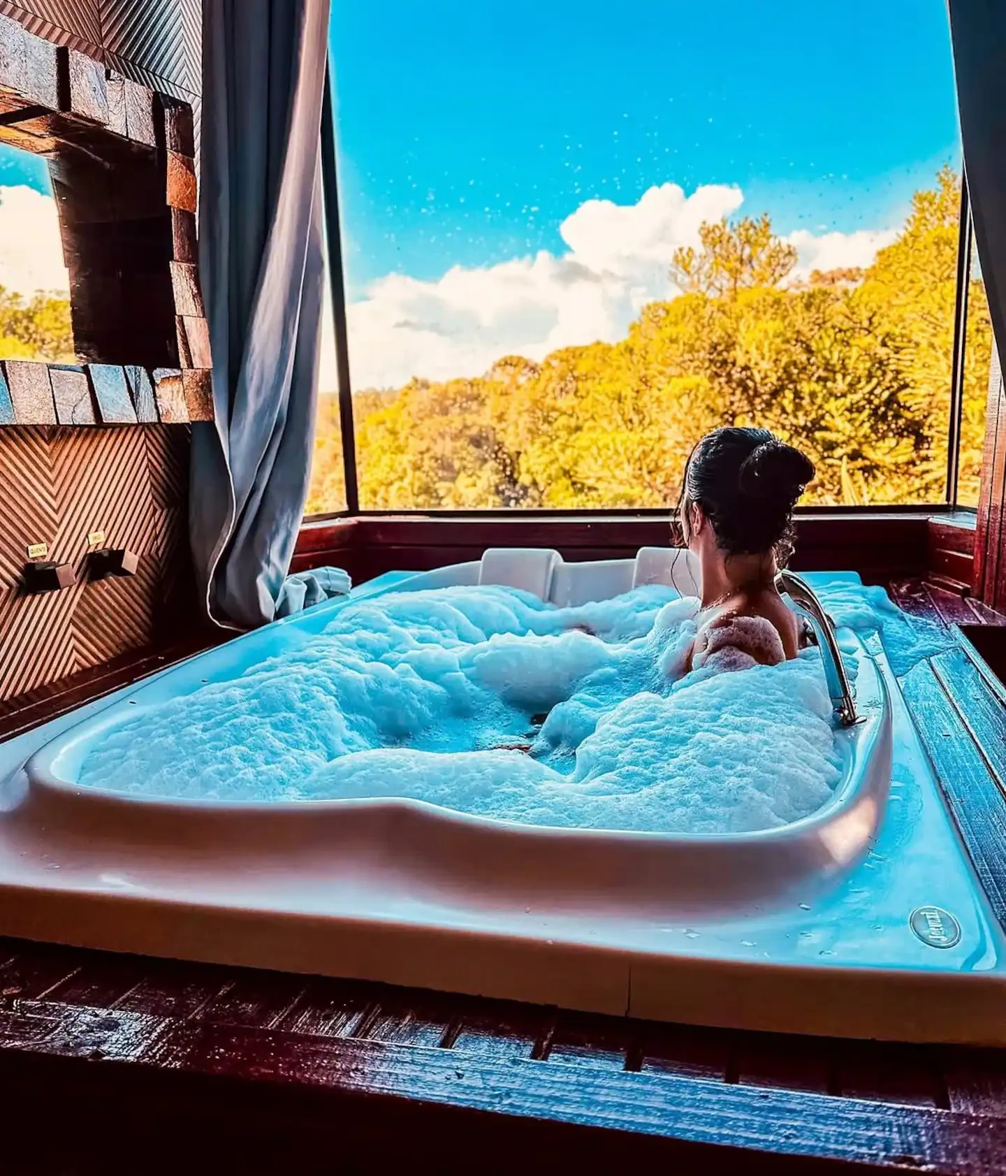 Hóspede relaxando na hidromassagem com espuma, diante de uma janela com vista para a mata de araucárias