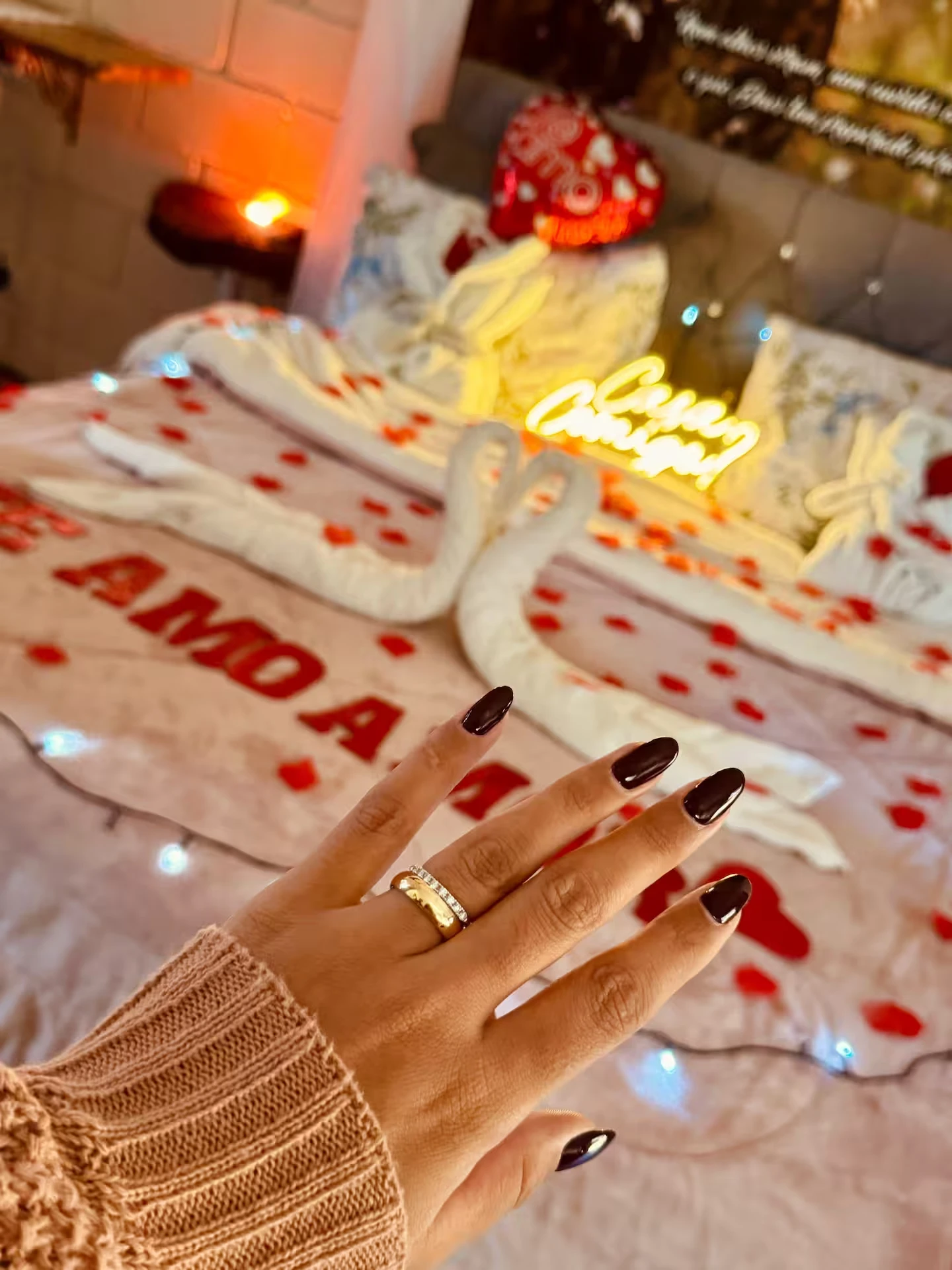 Mão com anel de noivado diante de uma cama decorada com pétalas e letreiro neon