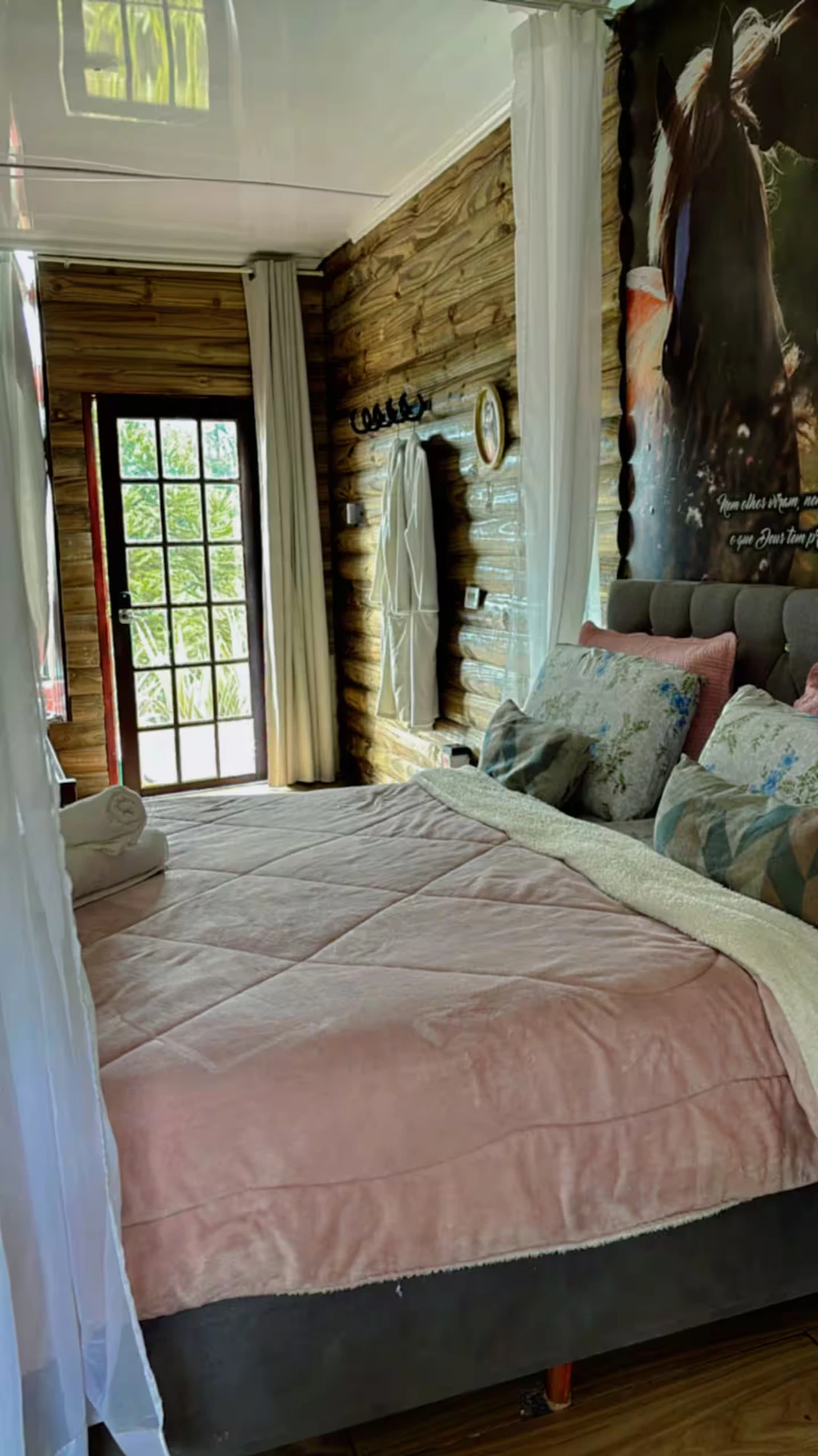 Quarto com cama de casal, edredom rosa, paredes de madeira e porta de vidro para o jardim