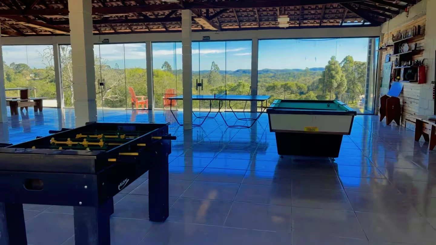 Salão com pebolim, mesa de pingue-pongue e sinuca, com paredes de vidro para a mata